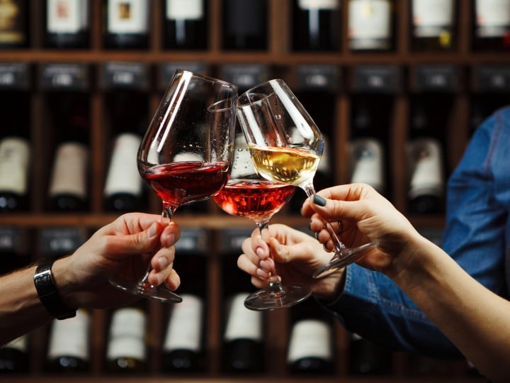 People in good mood clink glasses, celebrating together, photo focus on hands woman and man tasting degustation different types of wine, alcohol beverage to relax, bottles on shelves on background