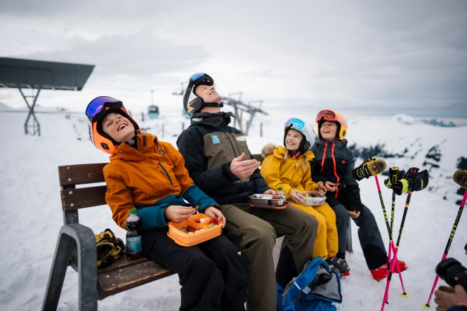 Skiass Hannes Reichelt sitzt mit Kindern auf einer Bank auf der Skipiste und alle lachen