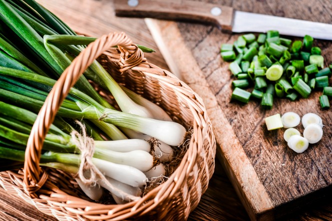 Young onion in wicker basket and chopped pieces with knife on wooden board in background..