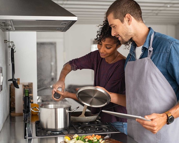 Frau und Mann kochen in einer Küche. Frau würzt das Gericht und der Mann schaut zu