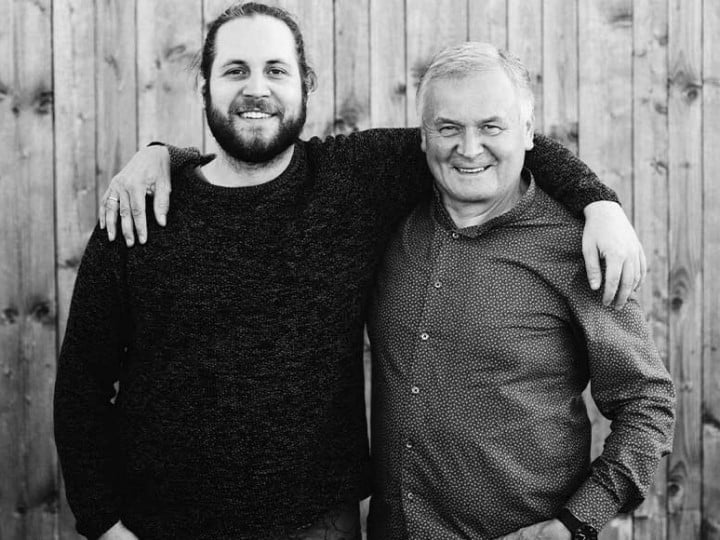 Vater und Sohn Gölles stehen vor einer Holzwand uns legen jeweils den Arm umeinenader, lächeln dabei in die Kamer