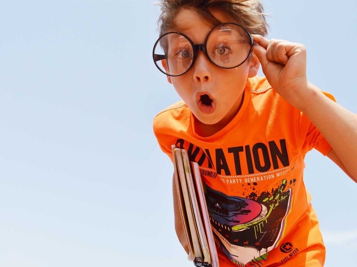 Schuljunge mit Brille, orangenem T-Shirt und Büchern unter seinem Arm schaut interessiert in die Kamera