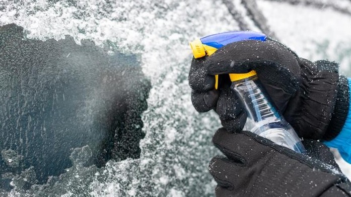 Vereiste Autoscheibe wird mit einem Enteiser-Spray eingesptüht. Wintereinbruch mit Eisregen. Hände in Winterhandschuhen und Enteiser Sprühflasche. 