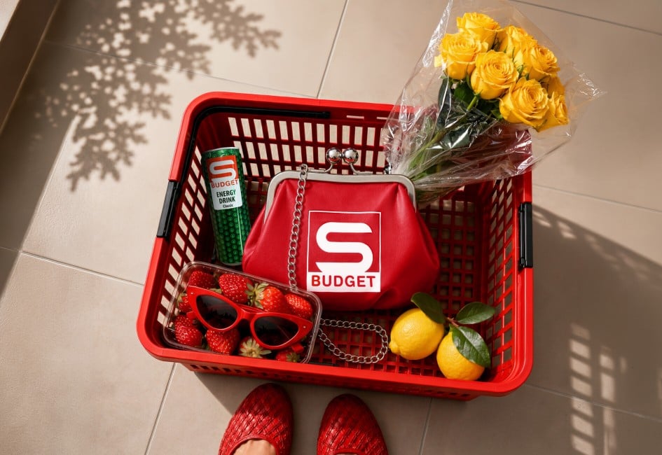 Die Börserl Bag liegt in einem roten Einkaufskorb, umgeben von anderen Produkten wie dem S-BUDGET Energy Drink, zwei Zitronen, einem Strauß gelber Rosen und einer Packung Erdbeeren. In den Erdbeeren liegt noch eine rote Sonnenbrille. Der Blick ist von oben auf den Korb gerichtet und der Korb steht auf einem neutralen Fliesenboden.
