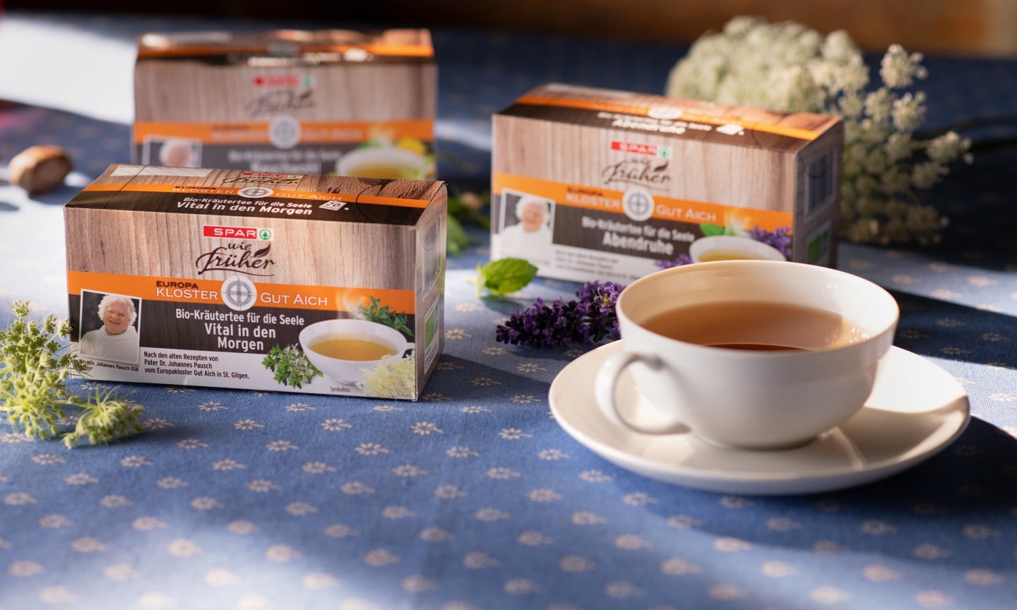 Das Bild zeigt drei Packungen „SPAR wie früher“ Bio-Kräutertees auf einem gedeckten Tisch mit einer Tasse Tee