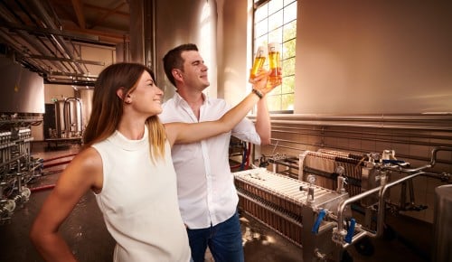 Geschäftsführer Markus Sautner und seine Frau, stehen in einer modernen Brauerei und halten jeweils ein Glas Bier in die Luft. 