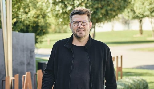 Hannes Reeh mit einer Weinflasche in der Hand im Hintergrund eine Gartenlandschaft