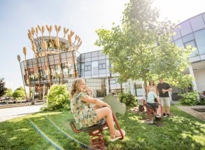 Außenansicht Haubiversum mit Spielplatz und Kindern auf einer Wiese vor dem Gebäude an einem sonnigen Tag