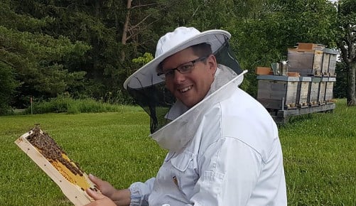 Manuel Petzl sitzend im Imkerkostüm mit einer Wabe in der Hand auf einer Wiese und im Hintergrund befinden sich Bienenstöcke
