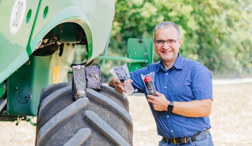 René Robitsch mit vier Wiener Gusto Produkten posiert bei einem Traktorfreifen im Hintergrund eine Ackerfläche mit Büschen