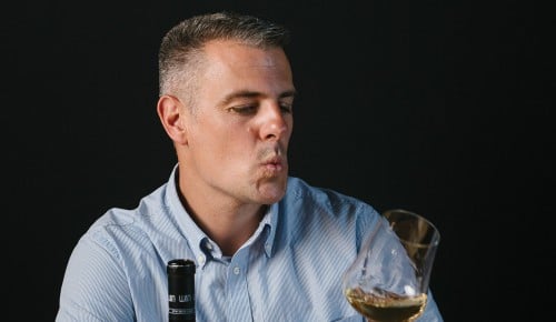 Kellermeister Georg Königsbauer verkostet gerade einen Weißwein auf einem Holztisch bei schwarzem Hintergrund