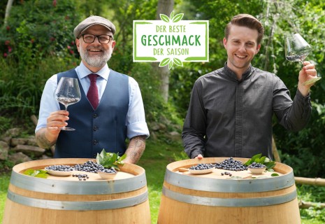 Hier sieht man zwei Sommeliers die in die Kamera lachen und ein Weinglas voll mit Heidelbeeren in der Hand haben.