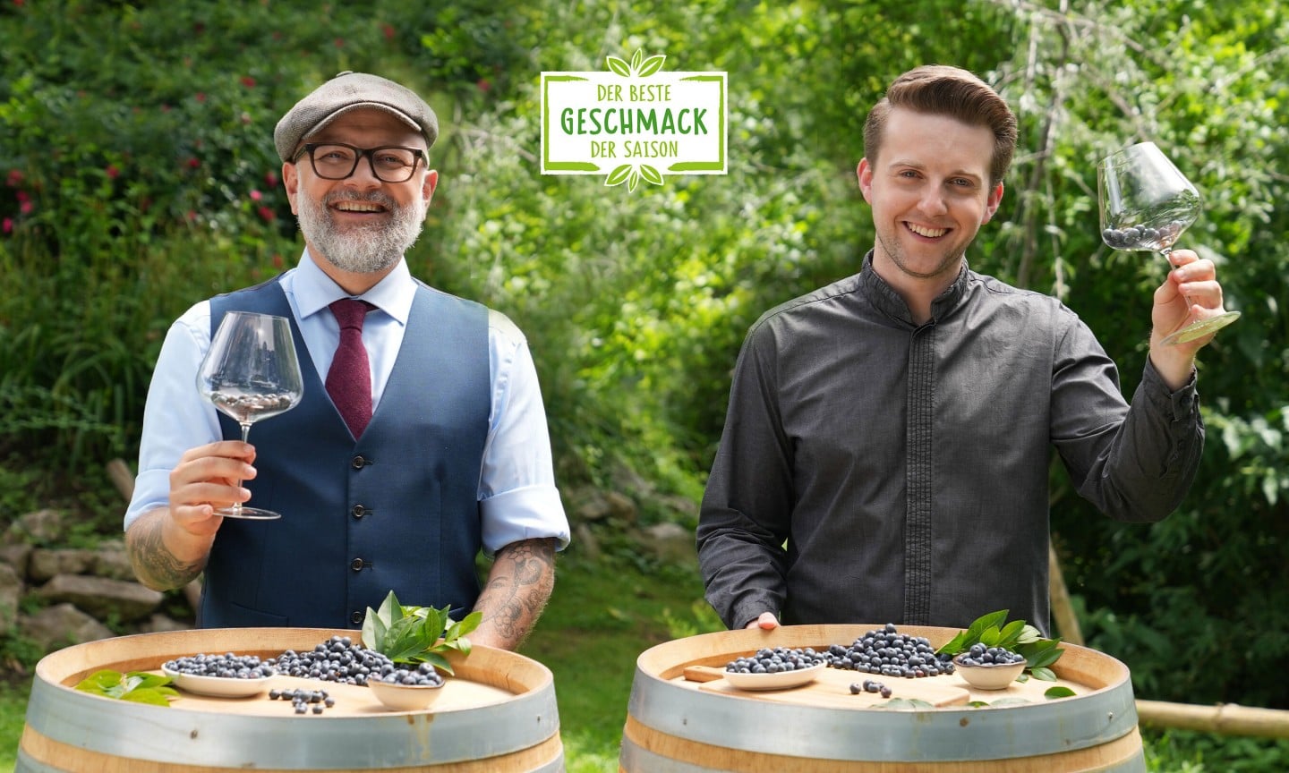 Hier sieht man zwei Sommeliers die beide Weingeläser gefüllt mit Heidelbeeren in die Luft halten und in die Kamera grinsen