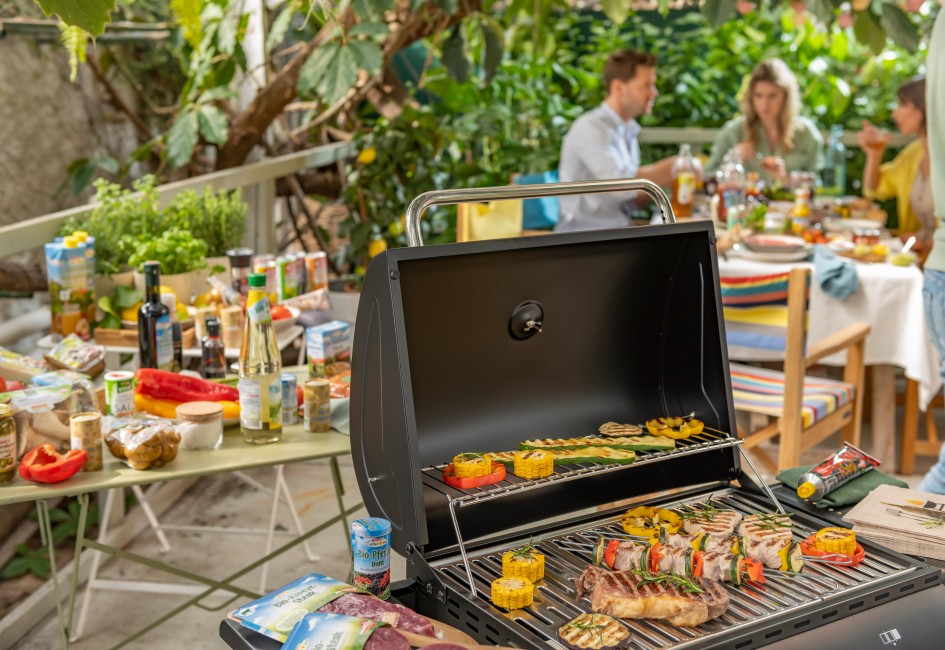 Grillfeier mit Freunden, ein Griller steht im Vordergrunf auf dem ein Steak und Gemüse gegrillt wird. Im Hintergrund sieht man diverse SPAR Produkte
