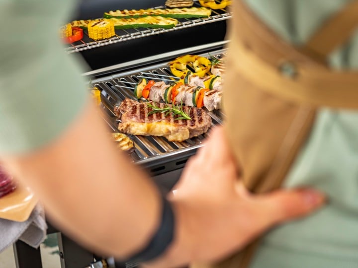 Man sieht einen Mann von hinten der vor einem Griller steht, auf dem Steaks Mais liegen