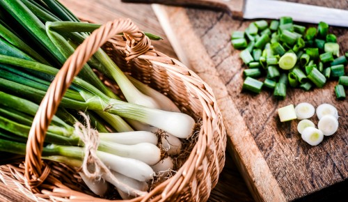 Young onion in wicker basket and chopped pieces with knife on wooden board in background..