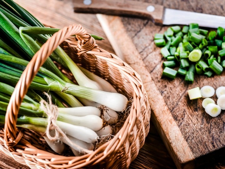 Young onion in wicker basket and chopped pieces with knife on wooden board in background..