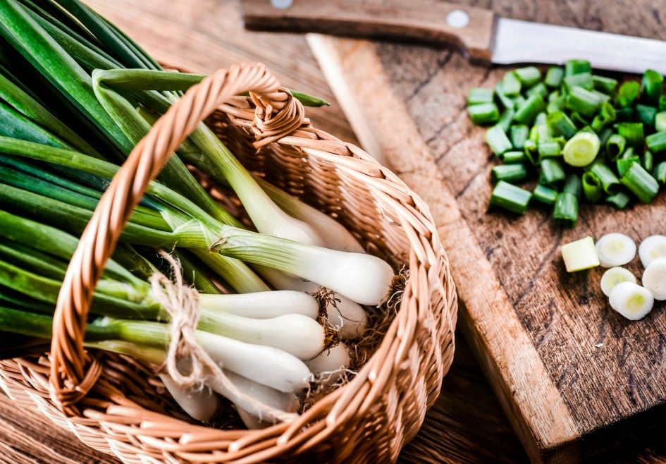 Young onion in wicker basket and chopped pieces with knife on wooden board in background..