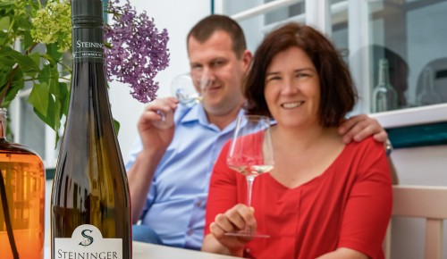 Eva und Peter Steininger von Weingut Steininger auf einer Holzbank mit Weingläsern in den Händen vor einem Haus