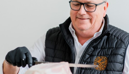 Mann mit weißem Hemd und schwarzer Giletjacke schneides mit einem Messer und schwarzen Handschuhen Schinken
